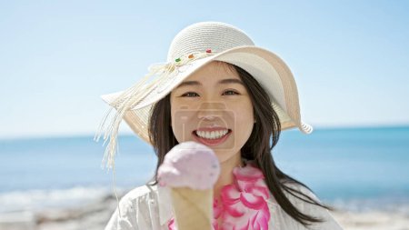 Téléchargez les photos : Jeune femme chinoise touriste souriant confiance manger de la crème glacée au bord de la mer - en image libre de droit