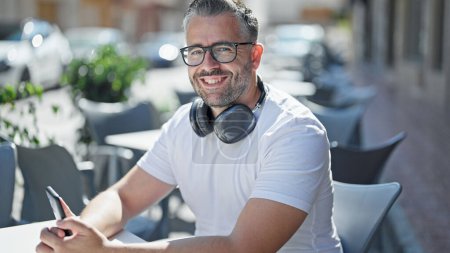 Téléchargez les photos : Homme aux cheveux gris utilisant un smartphone portant des écouteurs sur la terrasse du café - en image libre de droit