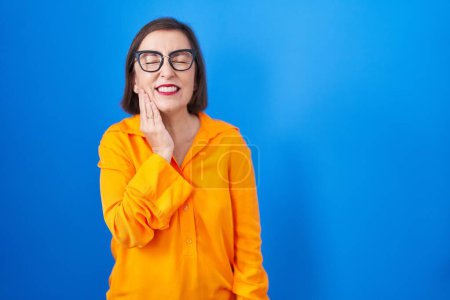 Photo for Middle age hispanic woman wearing glasses standing over blue background touching mouth with hand with painful expression because of toothache or dental illness on teeth. dentist - Royalty Free Image