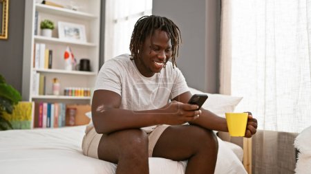 Foto de Hombre afroamericano usando smartphone bebiendo café en el dormitorio - Imagen libre de derechos