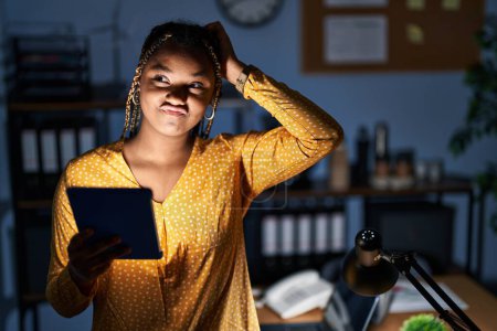 Téléchargez les photos : Femme afro-américaine avec des tresses travaillant au bureau la nuit avec tablette confondre et se poser des questions. incertain avec doute, pensant la main sur la tête. concept coûteux. - en image libre de droit