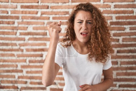 Photo for Young caucasian woman standing over bricks wall background angry and mad raising fist frustrated and furious while shouting with anger. rage and aggressive concept. - Royalty Free Image