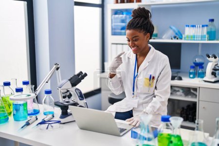 Téléchargez les photos : Jeune afro-américain avec des tresses travaillant au laboratoire scientifique avec ordinateur portable souriant heureux pointant avec la main et le doigt - en image libre de droit