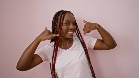 Photo for Cheerful african american woman with braids expressing confidence, performing a phone call gesture with hand over isolated pink background, captivating fun-loving joyful expression - Royalty Free Image
