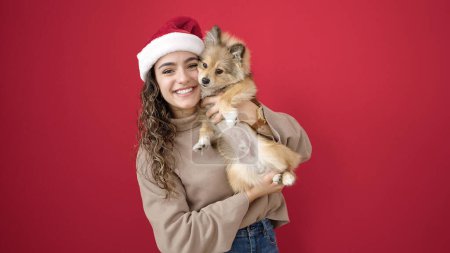 Téléchargez les photos : Jeune femme hispanique avec chien souriant confiant portant un chapeau de Noël sur fond rouge isolé - en image libre de droit