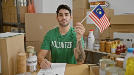 Téléchargez les photos : Jeune homme hispanique volontaire tenant le drapeau de la Malaisie au centre de charité - en image libre de droit