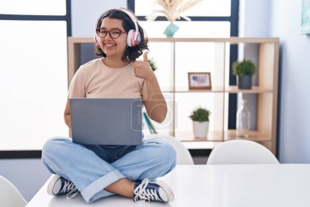 Photo for Young hispanic woman using laptop sitting on the table wearing headphones doing happy thumbs up gesture with hand. approving expression looking at the camera showing success. - Royalty Free Image