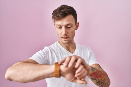 Photo for Caucasian man standing over pink background checking the time on wrist watch, relaxed and confident - Royalty Free Image