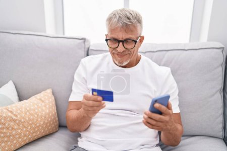 Téléchargez les photos : Homme aux cheveux gris du Moyen Âge utilisant smartphone et carte de crédit assis sur le canapé à la maison - en image libre de droit