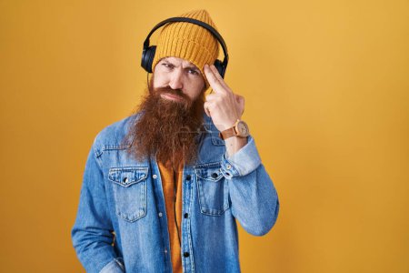 Foto de Hombre caucásico con barba larga escuchando música usando audífonos disparándose y matándose apuntando con la mano y los dedos a la cabeza como arma, gesto suicida. - Imagen libre de derechos