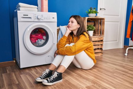 Photo for Young blonde woman sitting on floor waiting for washing machine at laundry room - Royalty Free Image