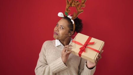 Foto de Mujer afroamericana escuchando sonido regalo de Navidad con orejas de reno sobre fondo rojo aislado - Imagen libre de derechos