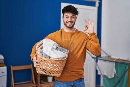 Foto de Hombre hispano con barba sosteniendo la cesta de la ropa en la lavandería haciendo signo bien con los dedos, sonriendo gesto amistoso excelente símbolo - Imagen libre de derechos