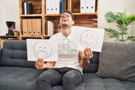 Photo for Young hispanic man working on depression holding sad to happy emotion paper smiling and laughing hard out loud because funny crazy joke. - Royalty Free Image