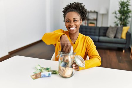 Téléchargez les photos : Femme afro-américaine assise sur la table tenant des billets de banque de bocal de charité à la maison - en image libre de droit