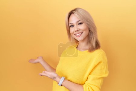 Foto de Mujer joven caucásica con suéter amarillo invitando a entrar sonriente natural con la mano abierta - Imagen libre de derechos