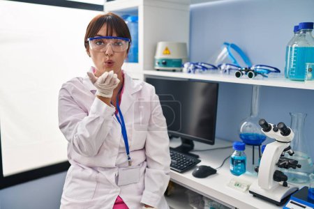 Foto de Young brunette woman working at scientist laboratory looking at the camera blowing a kiss with hand on air being lovely and sexy. love expression. - Imagen libre de derechos