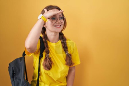 Photo for Young caucasian woman wearing student backpack over yellow background very happy and smiling looking far away with hand over head. searching concept. - Royalty Free Image