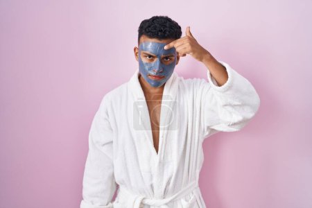Téléchargez les photos : Jeune homme hispanique portant un masque de beauté et peignoir de bain pointant malheureux à boutons sur le front, infection laide de la tête noire. acné et problème de peau - en image libre de droit