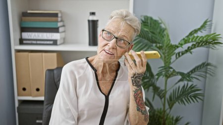 Téléchargez les photos : Femme âgée aux cheveux gris dans l'agitation des affaires, écoutant un message vocal raffiné sur son smartphone tout en travaillant au bureau - en image libre de droit