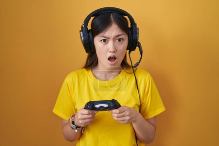 Téléchargez les photos : Jeune femme chinoise jouant contrôleur de tenue de jeu vidéo dans le visage de choc, regardant sceptique et sarcastique, surpris avec la bouche ouverte - en image libre de droit