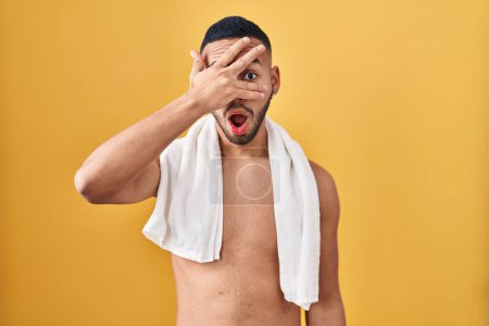 Foto de Joven hispano de pie sin camisa con toalla asomándose en shock cubriendo la cara y los ojos con la mano, mirando a través de los dedos con expresión avergonzada. - Imagen libre de derechos