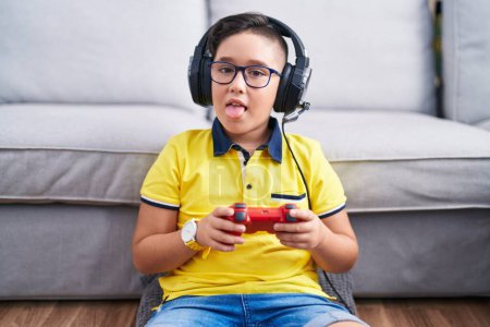 Foto de Joven niño hispano jugando videojuego sosteniendo controlador usando auriculares sacando la lengua feliz con expresión divertida. concepto de emoción. - Imagen libre de derechos
