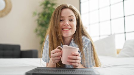 Foto de Mujer rubia joven haciendo videollamada con portátil bebiendo café en el dormitorio - Imagen libre de derechos