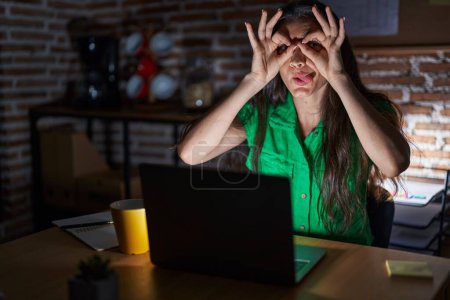 Photo for Young teenager girl working at the office at night doing ok gesture like binoculars sticking tongue out, eyes looking through fingers. crazy expression. - Royalty Free Image