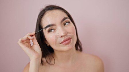 Téléchargez les photos : Jeune belle femme hispanique appliquant maquillage sur les sourcils sur fond rose isolé - en image libre de droit