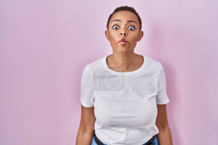 Foto de Hermosa mujer afroamericana de pie sobre fondo rosa haciendo cara de pez con labios, gesto loco y cómico. expresión divertida. - Imagen libre de derechos