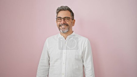Foto de Hombre hispano de pelo gris, seguro, con una sonrisa encantadora, gafas, de pie sobre un fondo rosa aislado, radiando positividad - Imagen libre de derechos