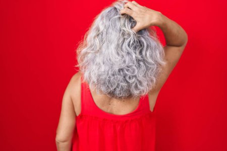 Foto de Mujer de mediana edad con el pelo gris de pie sobre fondo rojo hacia atrás pensando en la duda con la mano en la cabeza - Imagen libre de derechos