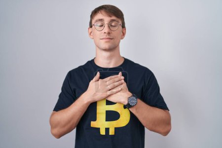Téléchargez les photos : Homme blond caucasien portant Bitcoin t-shirt souriant avec les mains sur la poitrine avec les yeux fermés et geste reconnaissant sur le visage. concept de santé. - en image libre de droit
