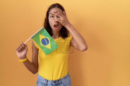 Téléchargez les photos : Jeune femme hispanique tenant le drapeau brésilien jetant un coup d'oeil dans le visage de couverture de choc et les yeux avec la main, regardant à travers les doigts avec une expression embarrassée. - en image libre de droit