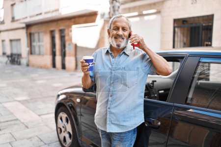 Téléchargez les photos : Senior grey-haired man talking on smartphone drinking coffee standing by car at street - en image libre de droit