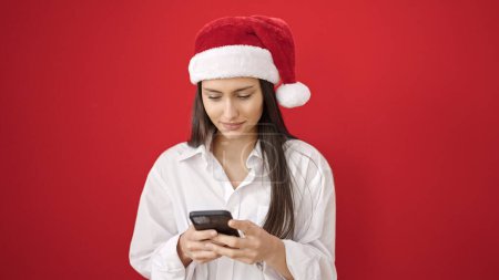 Téléchargez les photos : Jeune belle femme hispanique portant un chapeau de Noël en utilisant un smartphone sur fond rouge isolé - en image libre de droit