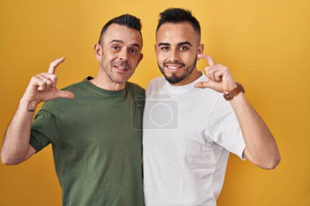 Téléchargez les photos : Couple homosexuel debout sur fond jaune geste souriant et confiant avec la main faisant signe de petite taille avec les doigts à la recherche et la caméra. concept de mesure. - en image libre de droit
