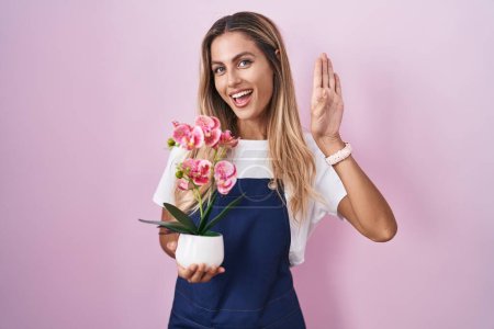 Foto de Joven mujer rubia vistiendo delantal jardinero sosteniendo planta renunciando a decir hola feliz y sonriente, gesto de bienvenida amistoso - Imagen libre de derechos