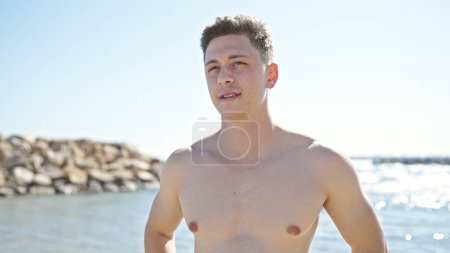 Téléchargez les photos : Jeune homme hispanique debout touristique avec une expression détendue au bord de la mer - en image libre de droit