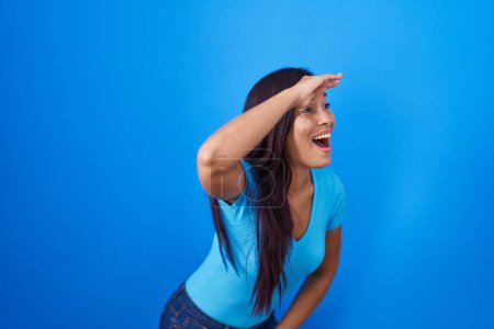 Photo for Young hispanic woman standing over blue background very happy and smiling looking far away with hand over head. searching concept. - Royalty Free Image