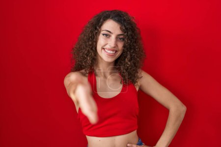 Photo for Hispanic woman with curly hair standing over red background smiling friendly offering handshake as greeting and welcoming. successful business. - Royalty Free Image