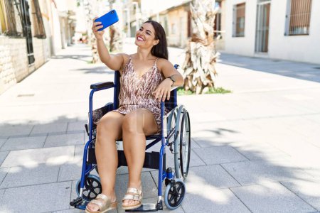Téléchargez les photos : Jeune belle femme hispanique faire selfie par smartphone assis sur fauteuil roulant à la rue - en image libre de droit