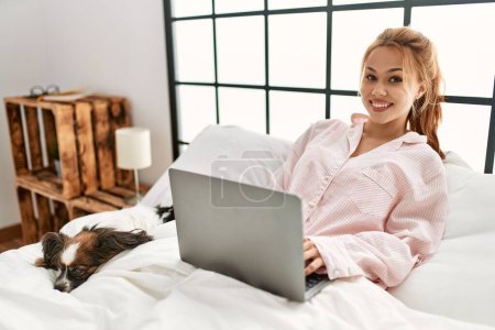 Téléchargez les photos : Jeune femme caucasienne utilisant ordinateur portable assis sur le lit avec chien à la chambre - en image libre de droit