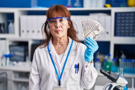 Foto de Mujer hispana de mediana edad que trabaja en un laboratorio científico sosteniendo una actitud de pensamiento de dólares y una expresión sobria que parece segura de sí misma - Imagen libre de derechos
