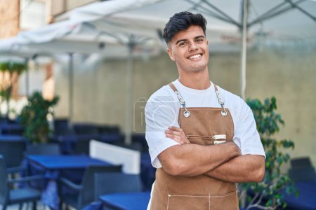 Téléchargez les photos : Jeune homme hispanique serveur souriant confiant debout avec les bras croisés geste à la terrasse du café - en image libre de droit