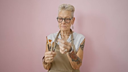 Téléchargez les photos : Une artiste senior aux cheveux gris au visage sérieux tenant intensément des pinceaux sur un fond rose isolé - une classe de peinture en plein essor - en image libre de droit