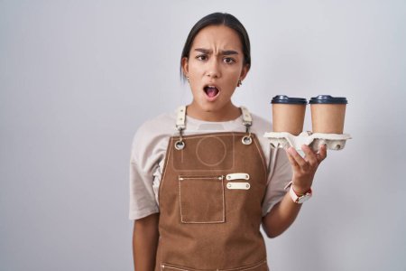 Photo for Young hispanic woman wearing professional waitress apron holding coffee in shock face, looking skeptical and sarcastic, surprised with open mouth - Royalty Free Image