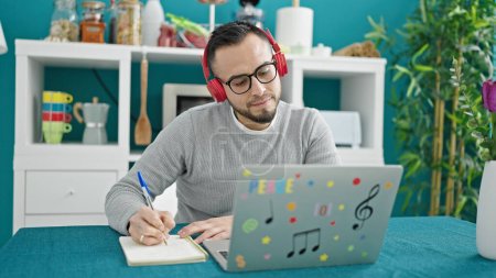 Foto de Hombre hispano usando auriculares usando computadora portátil escribiendo en un cuaderno en el comedor - Imagen libre de derechos