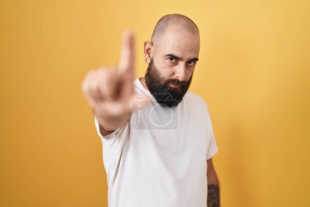 Foto de Joven hombre hispano con barba y tatuajes de pie sobre fondo amarillo señalando con el dedo hacia arriba y expresión enojada, sin mostrar ningún gesto - Imagen libre de derechos